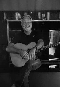 Denny Follis, musician; sits and smiles with his acoustic guitar